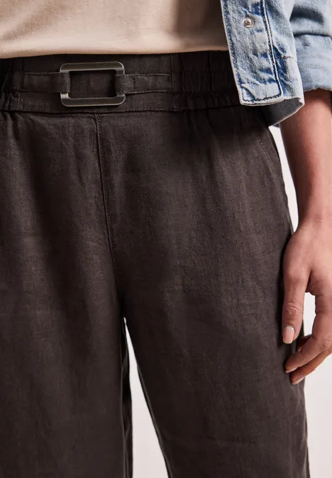 Schwarze Leinenhose mit breitem Bund und Gürtelschnalle, kombiniert mit Jeansjacke und einer Hand an der Hüfte.